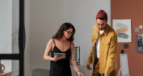 Two young professionals in an office, a woman holding a tablet and a man wearing a red beanie looking at a desk.